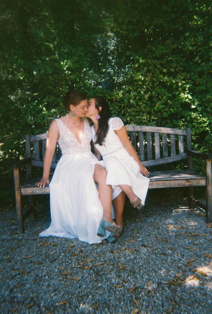 Deux mariées s'embrassent assises sur un banc. Amor amor - Photographies de mariage à l'argentique, par Sabine Greppo.